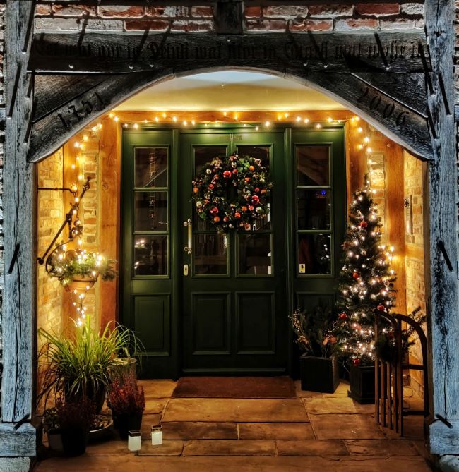 Festive cozy yet grand entrance to a home, green door with Christmas wreath bordered by white Christmas lights, candles on the flagstone floor and lit Christmas tree – inspiration for a weekly list of free educational events and resources for the association community